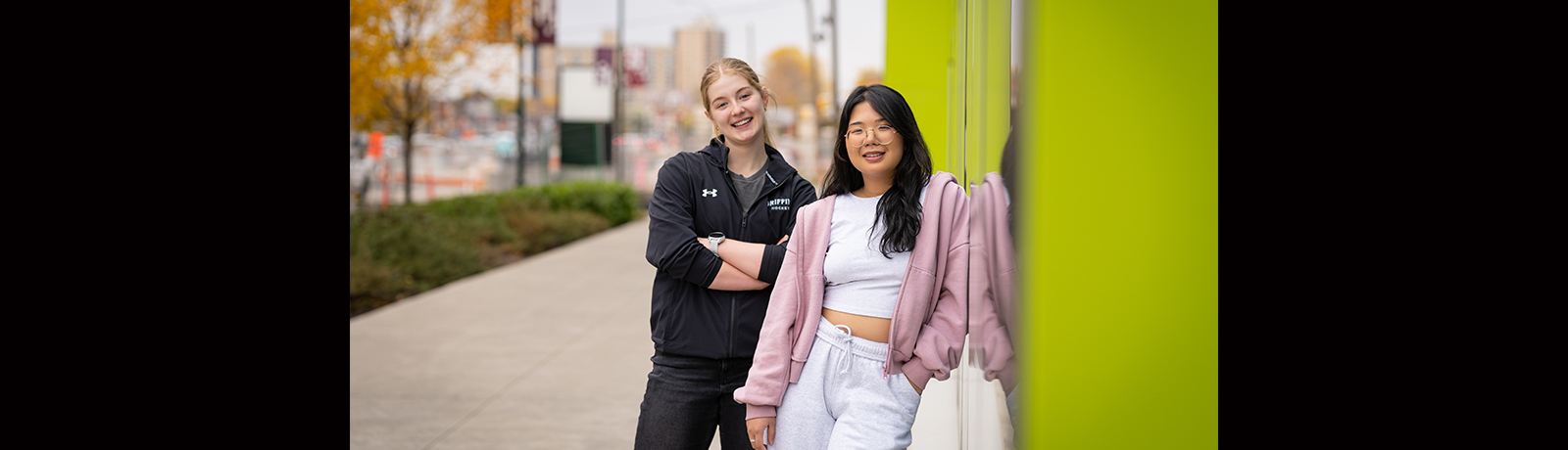 Two students stand outdoors, next to each other, one with arms crossed and one leaning against a building.