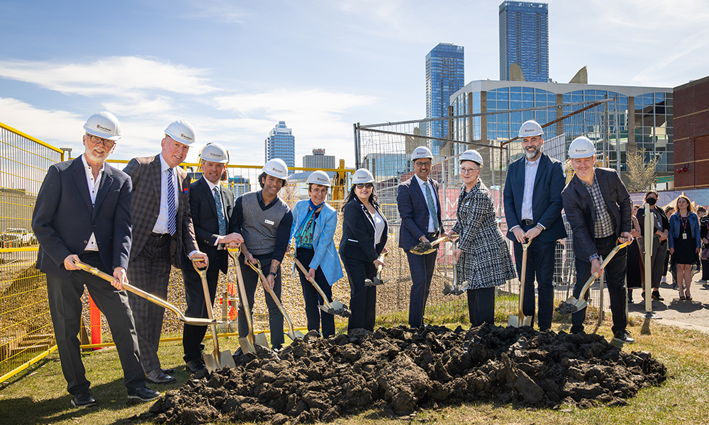 Several people wear hard hats and lift dirt on shovels at a groundbreaking ceremony.