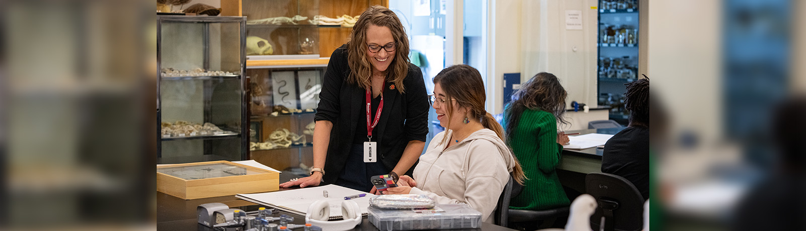 A teacher stands helping a student who sits at a table in a lab