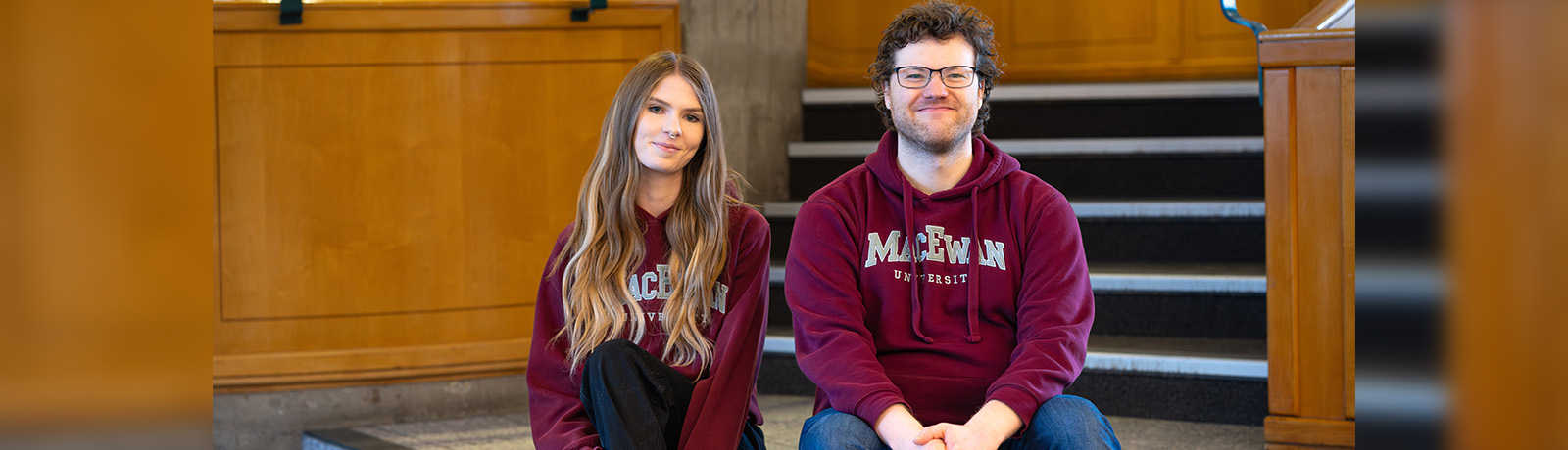 A young woman and young man sit on the tiled steps at the bottom of the spiral staircase, both wearing MacEwan hoodies.