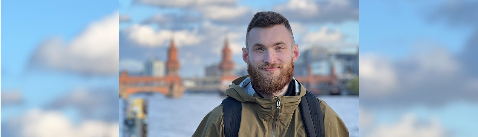 A man in a green jacket wearing a backpack stands outside, with a body of water and architectural buildings in the background