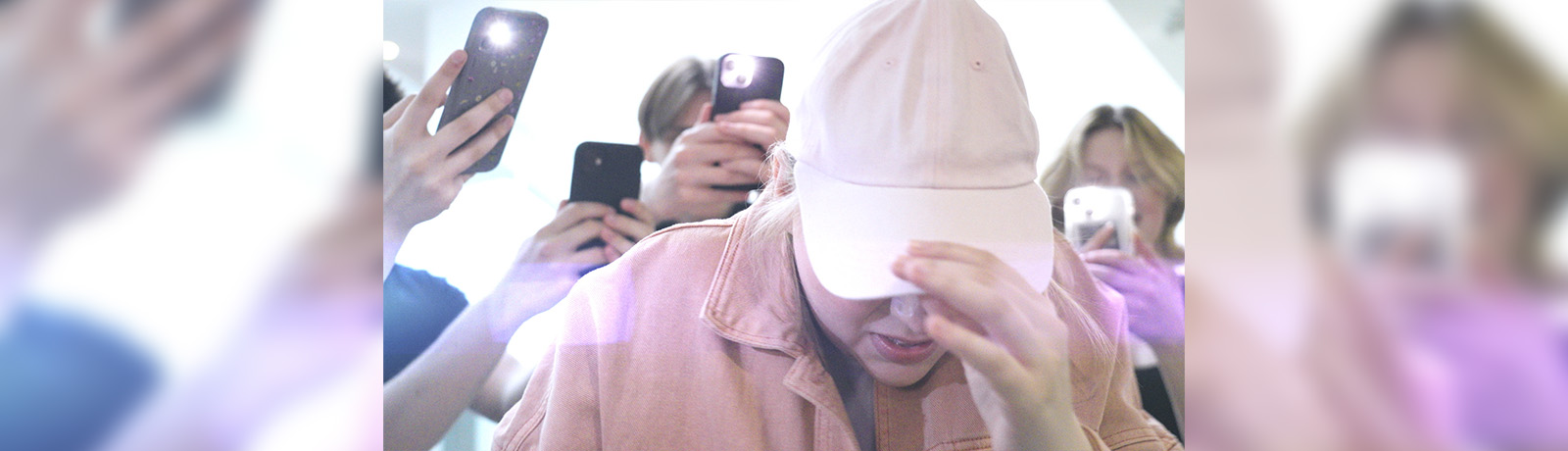 A female student pulls a white ball cap low over her face as other students record her on cell phones.
