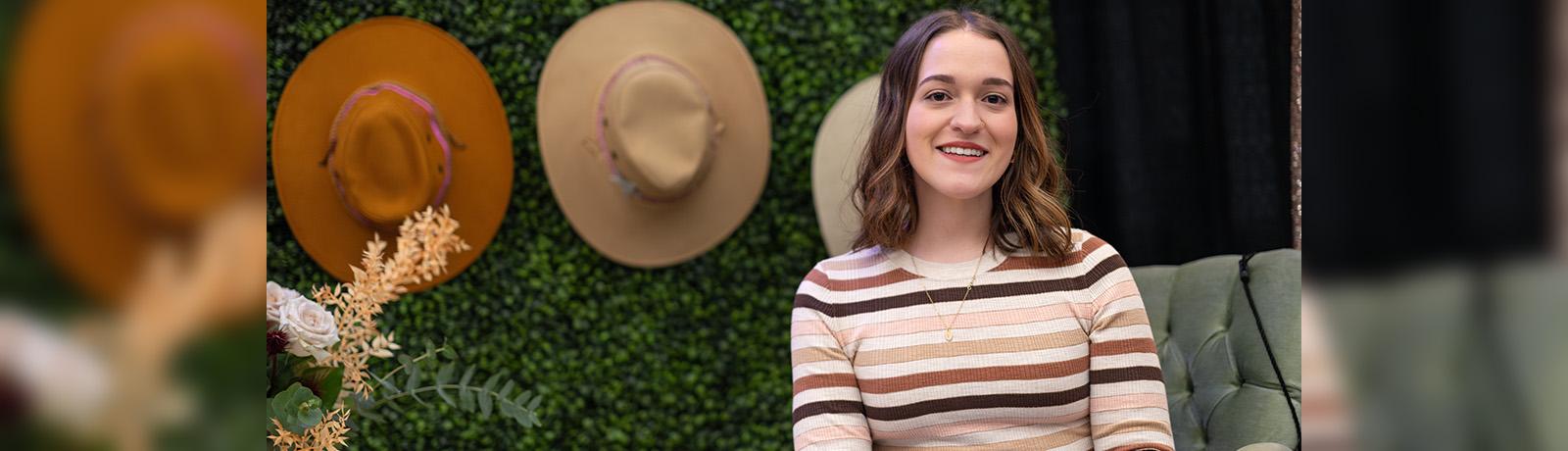 Sofia Fusco sits in front of a wall of green leaves with cowboy hats hanging on it decoratively.