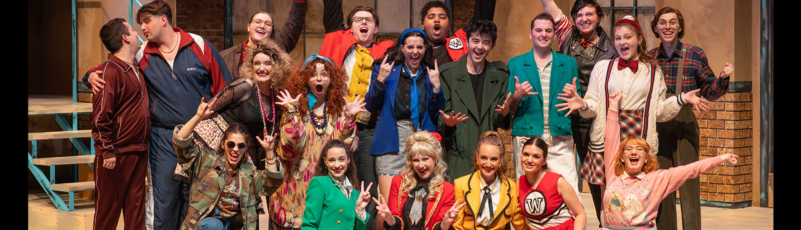 The cast of Heathers on stage with their hands in the air, dressed in colourful costumes