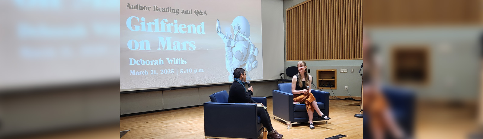 Two women sit in chairs angled toward one another on a stage, with the book cover of Girlfriend on Mars projected onto the wall behind them.