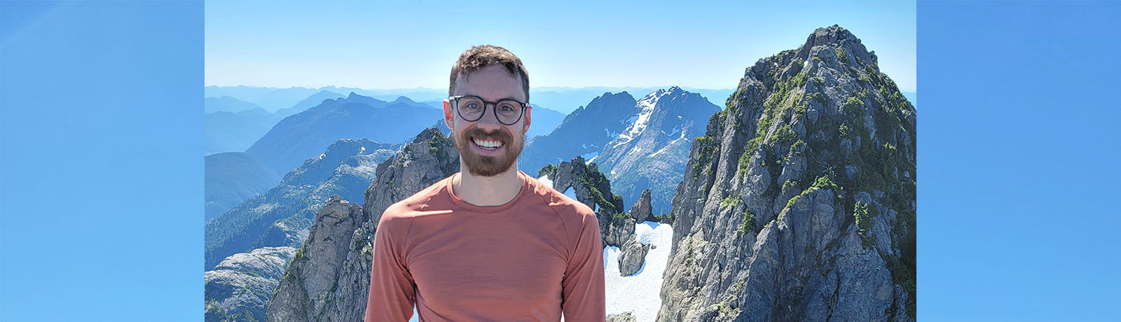 Jay Wickins smiles from the top of a mountain, with other rocky peaks behind him.