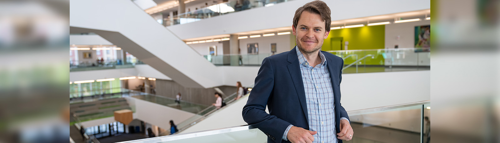 Dr. Aidan Forth wears a blue blazer and leans back against a glass railing in Allard Hall.