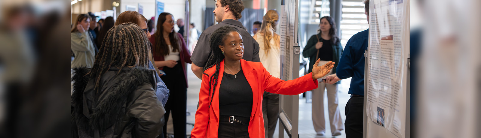 A student in a red blazer points to her poster project, explaining her research to two onlookers.