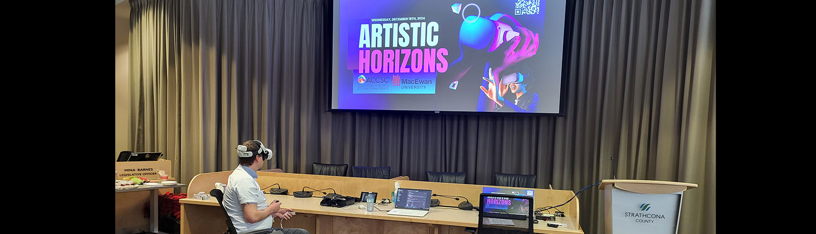 A student wears VR goggles and looks at a large screen on the wall with an image that says "Artistic Horizons"