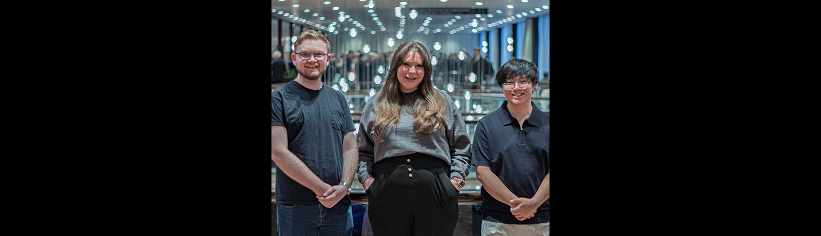 Aidan Lapp, Ashlee Rolheiser and Nathan Poon stand side-by-side in front of a glass railing, with lights hanging at various heights behind them.