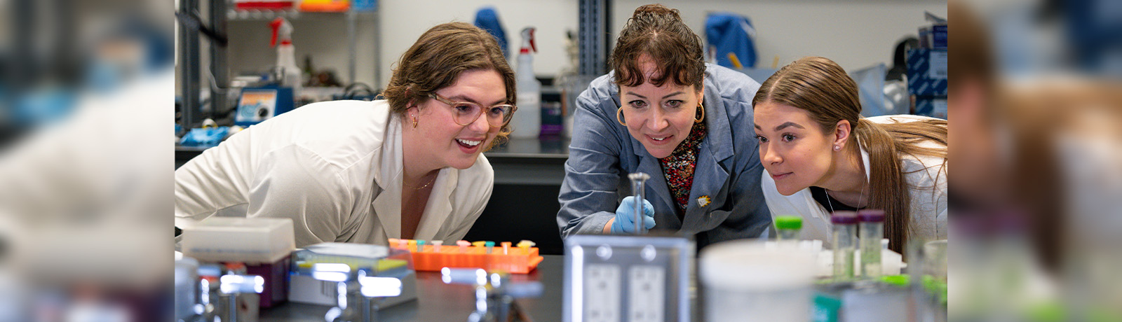 Three women in lab coats look at a test tube in a stand on a metal table in a lab setting.