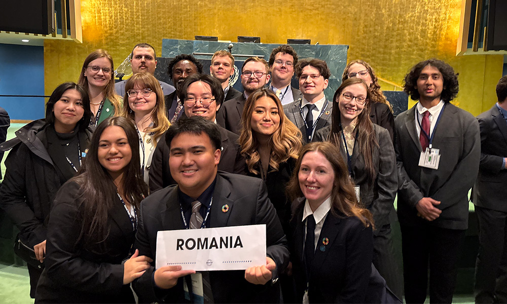 A large group of students pose together in front of a gold wall, holding a sign that says "Romania."