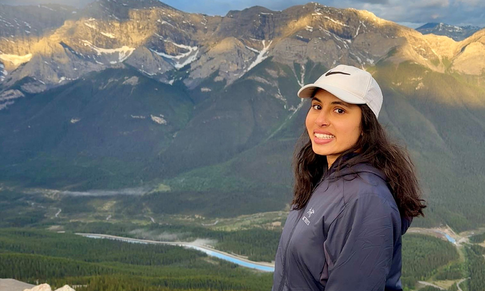 A woman stands in front of a background of mountains and a valley with a river
