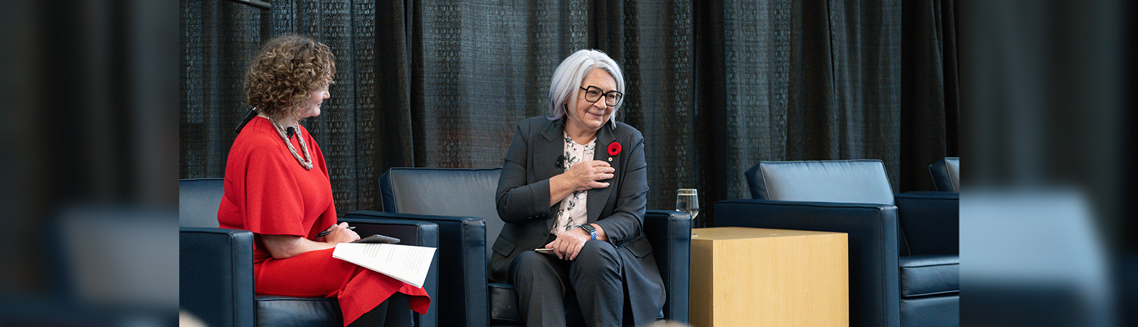 two women sit on stage in large grey chairs. One is looking toward the audience and speaking, and the other is looking at speaker