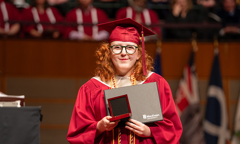 a woman stands on stage in a red graduation cap and gown holding a parchment