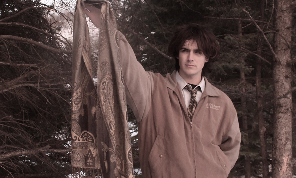 Sawyer Begg stands in front of a forested background holding a patterned scarf  in front of him
