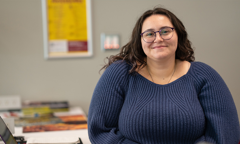 Leanna Bressan wears a blue sweater and smiles while sitting at a table on campus