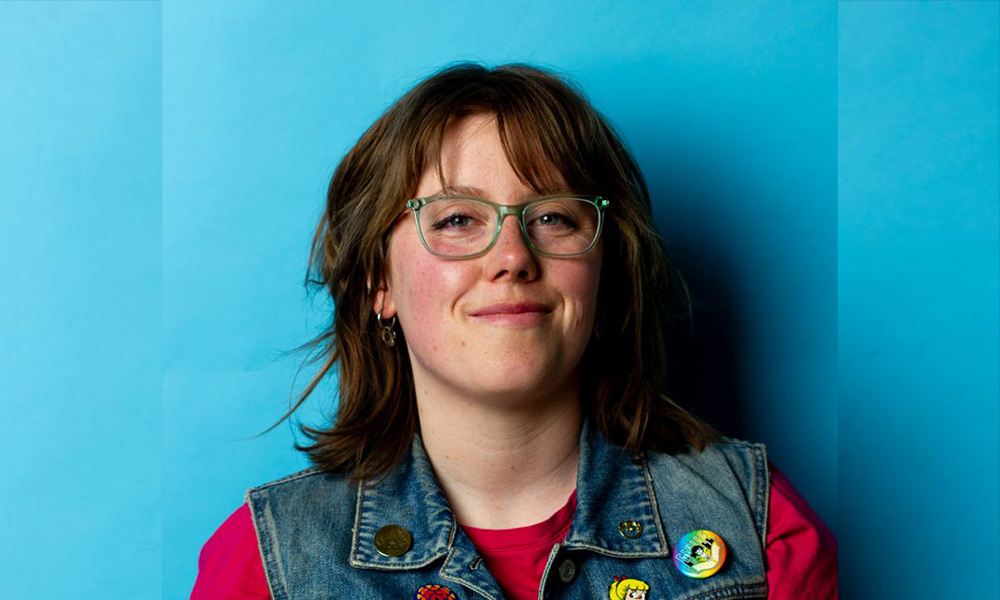 Quinlyn Hawkswell smiles, wearing a jean jacket and standing against a blue background