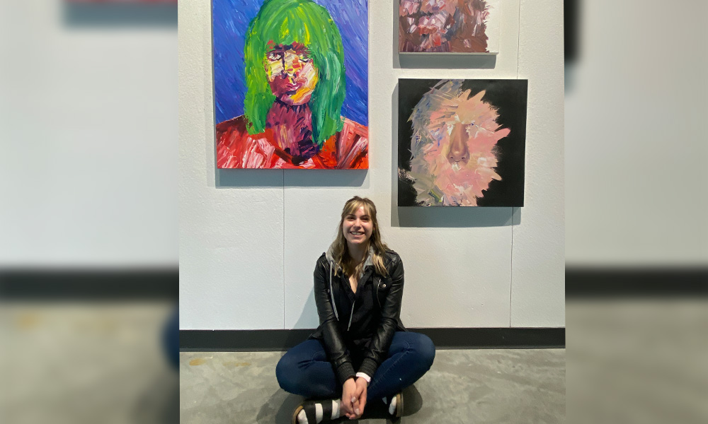 Natalie Potts sits on the floor in front of a wall that displays three paintings