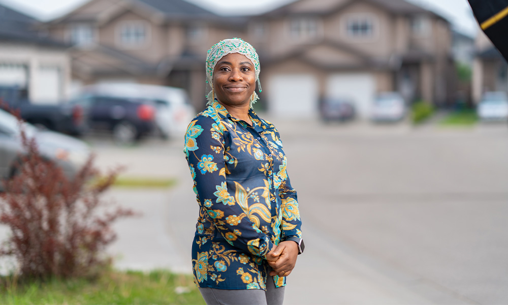 A woman stands in a driveway, smiling, hands held together in front of her.