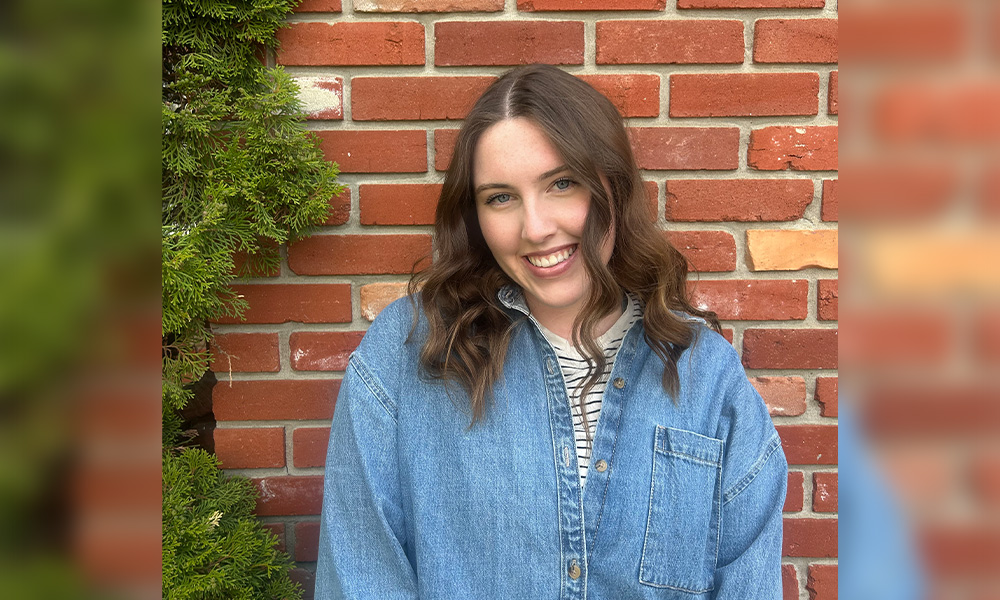 A woman in a denim shirt stands in front of a red brick wall.