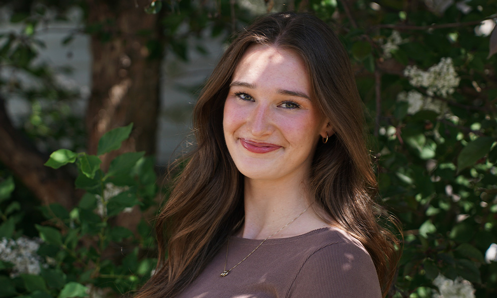 A woman smiles, standing in the shade of flowering trees.