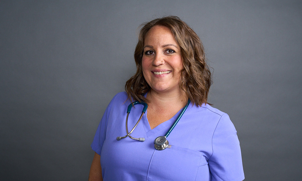 a woman standing smiling at the camera, in purple scrubs with a stethoscope around her neck