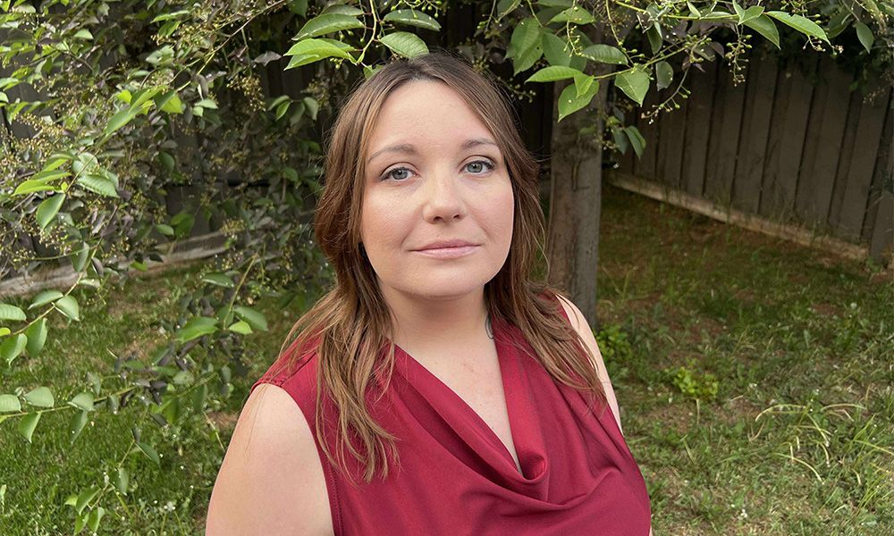 A woman stands in front of a lush, green tree, wearing a red sleeveless shirt with a cowl neck.