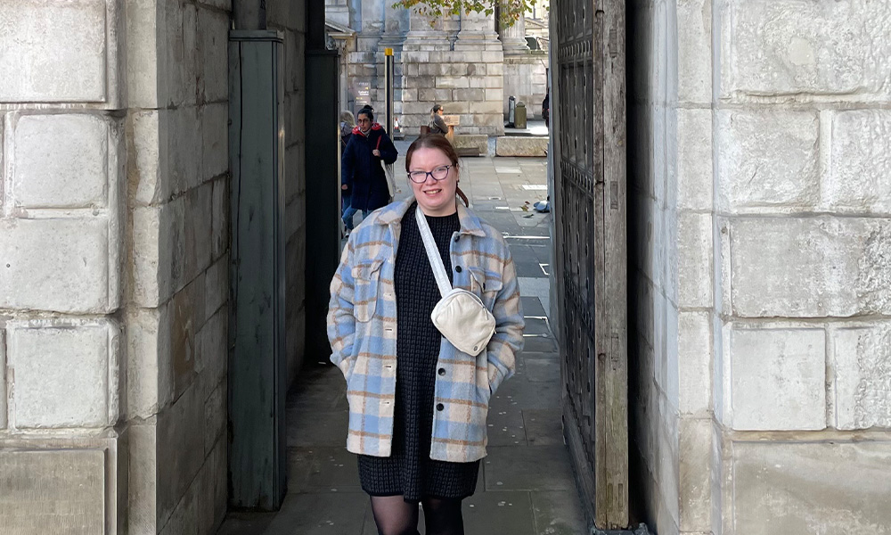 A woman in a plaid jacket stands in a stone archway in the middle of a city square.