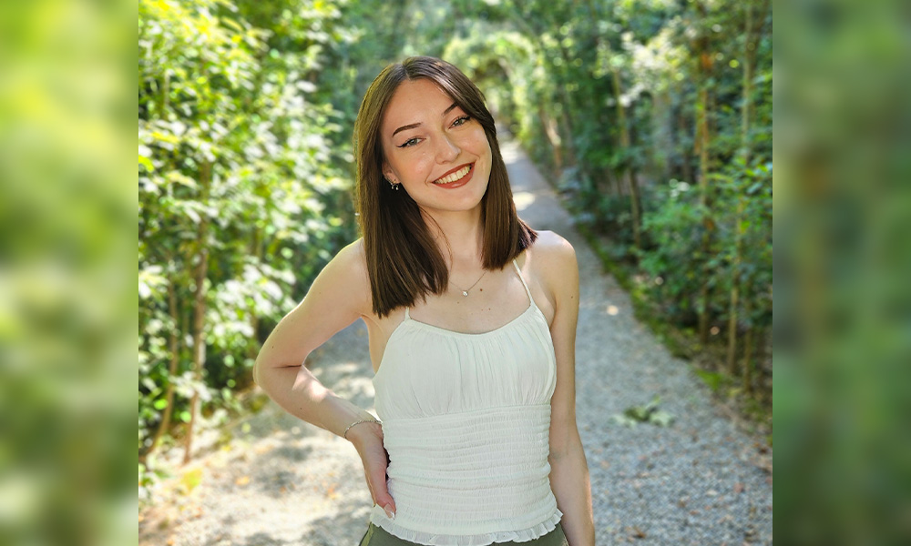 A woman wears a white tank top and stands on a hiking path, surrounded by full, green trees
