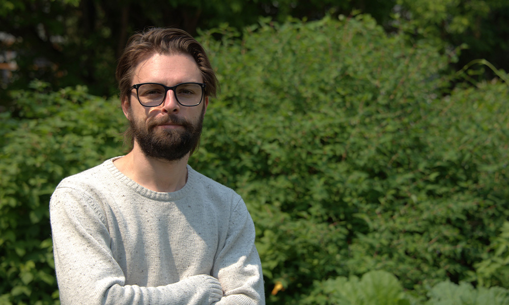 A man stands in front of green trees and folds his arms over his chest.