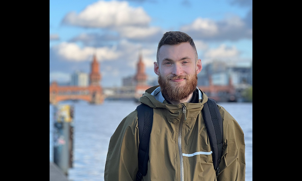 A man wears a green jacket and black backpack, with a river and city slightly blurred in the background.