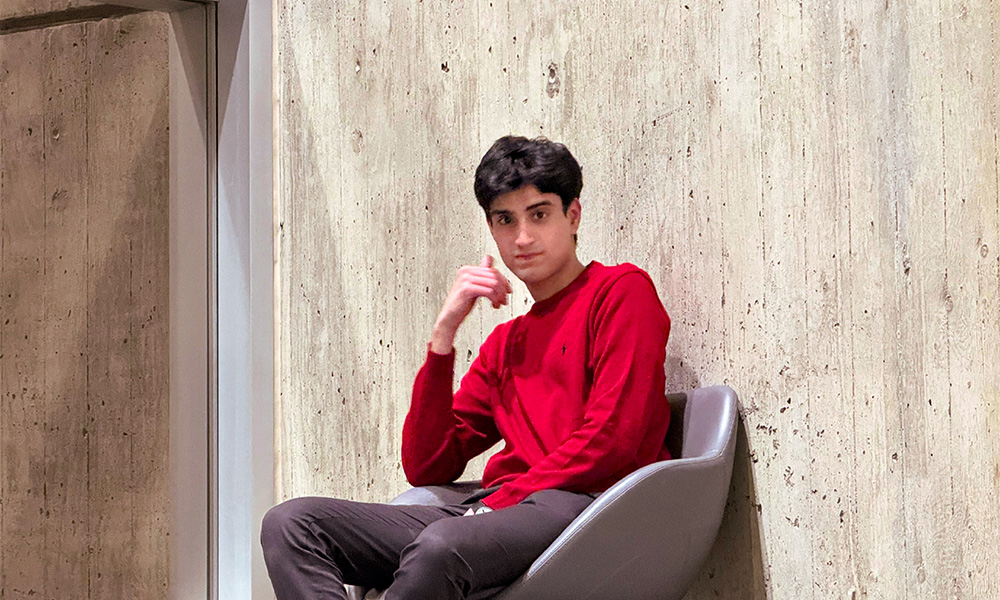 A man in a red shirt sits in a grey chair against a concrete wall.
