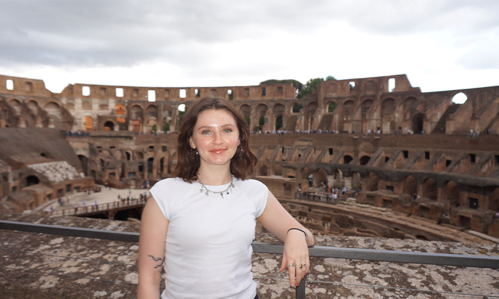 A woman in a white tee shirt leans against a metal railing, overlooking the interiror of the Roman Colosseum in the background.