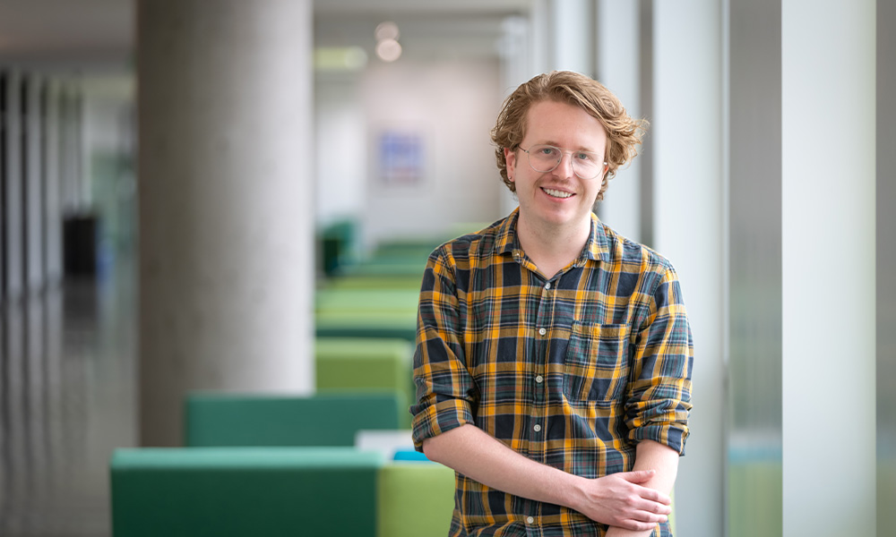 Matt Gaudet wears an orange plaid shirt and stands in front of green seating in Allard Hall.