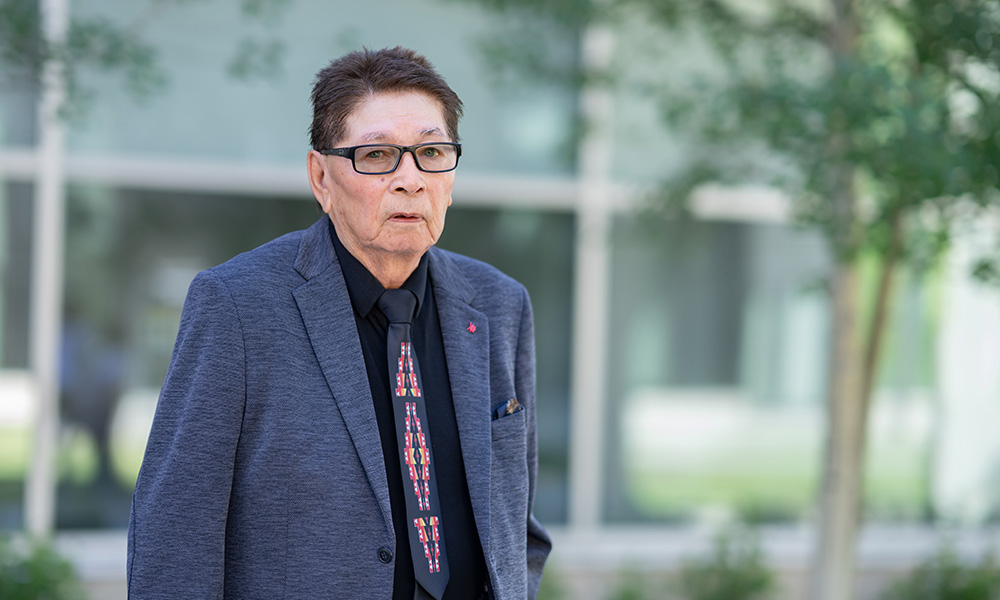 Bernie Makokis stands outside in front of a tree, wearing a blue suit