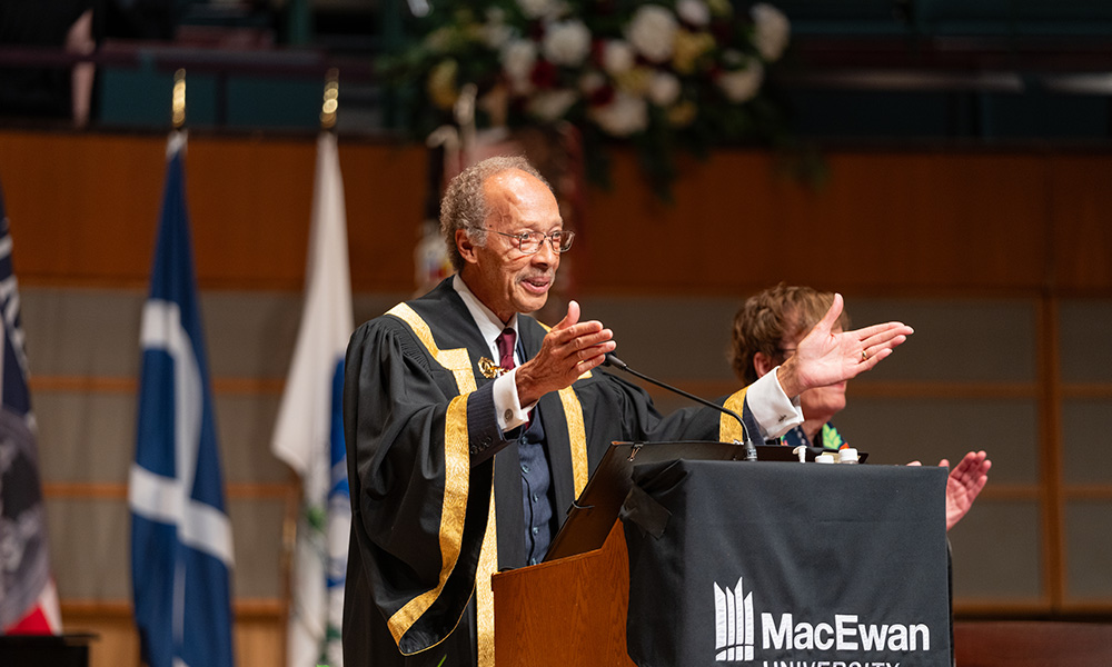 a man stands at a podium, in a graduation gown, speaking to an audience