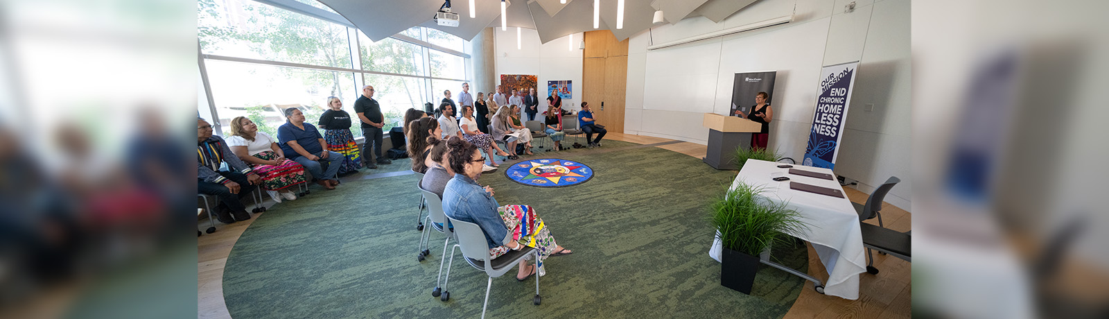 Many people sit in chairs in a semi-circle, looking to Dr. Trimbee addressing them at a podium.