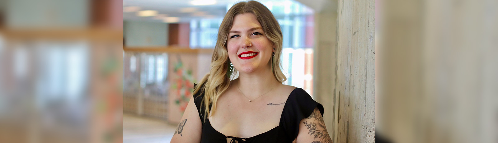 Sam Micka wears a black top and leans against a concrete pillar beside the spiral staircase on campus.