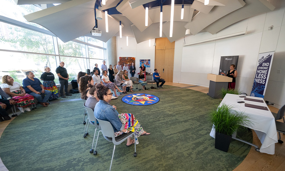 People sit in chairs in a semi-circle, looking to Dr. Trimbee who addresses them from behind a podium.