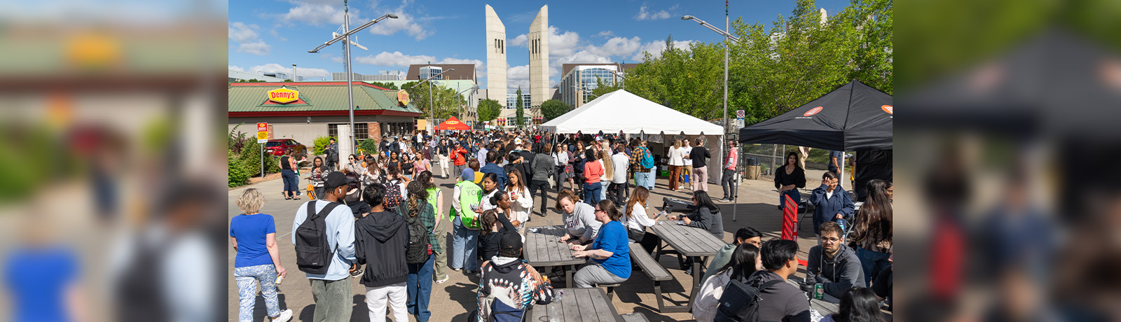 Students, staff and faculty walk down 108 Street and sit around picnic tables, with MacEwan's towers in the background.