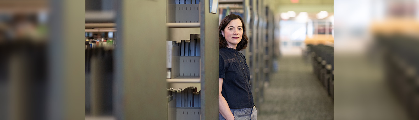 Beth Graham leans against a metal bookshelf in the MacEwan Library, wearing a dark tee shirt and jeans.