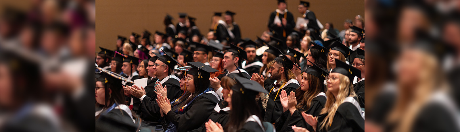 Graduating students in black caps and gowns sit in a theatre together, clapping