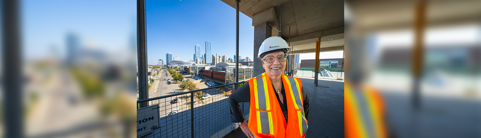 a woman in a hard hat and safety vest stands within a building under construction overlooking a busy downtown street