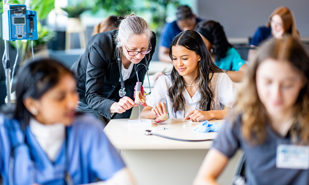 A teacher leans over a table to help a nursing student wearing scrubs with a problem.