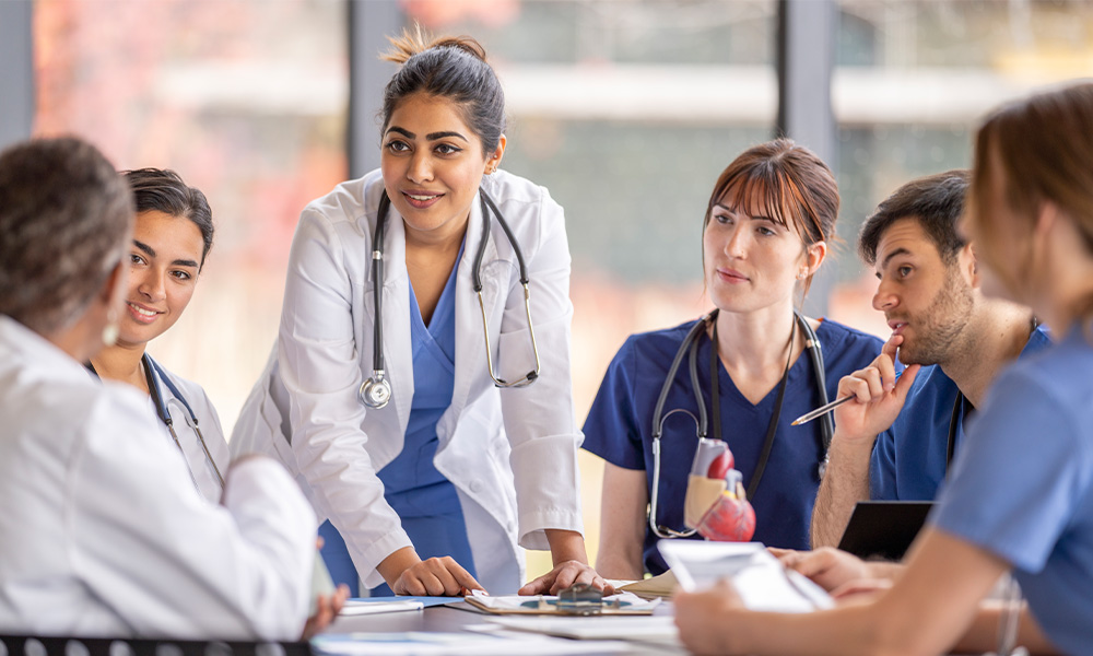 A nursing student stands at a table and speaks to other nursing students seated around it.