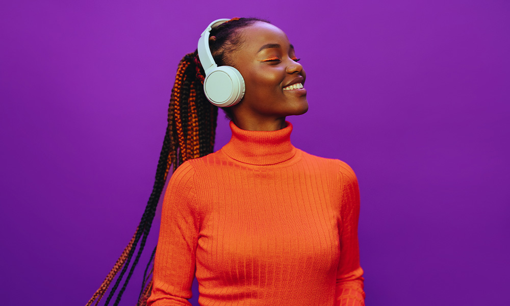 A woman wears an orange sweater and headphones, standing in front of a purple background.