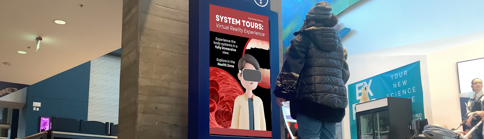 A person in a winter jacket stands in front of a display in the Telus World of Science that says System Tours