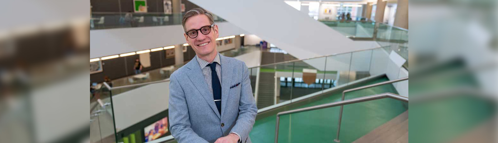 Dr. Lorkovic wears a grey suit and leans against a glass railing in Allard Hall.