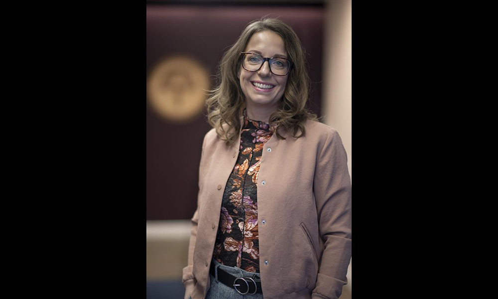 Ashley Baxter leans against a beige wall, wearing a brown blazer over a floral shirt.